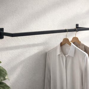 以下が含まれることがあります： ミニマルなデザインの黒い金属製の壁掛けハンガーラック。2つの木製ハンガーには、白いボタンダウンシャツとベージュのブレザーがかかっています。ラックは質感のある白い壁に取り付けられています。