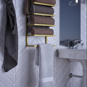May include: A gold-colored metal towel rack with five shelves, holding rolled brown towels. A gray towel hangs from the bottom. A dark gray towel hangs on the left. The rack is mounted on a white, chevron-patterned wall.