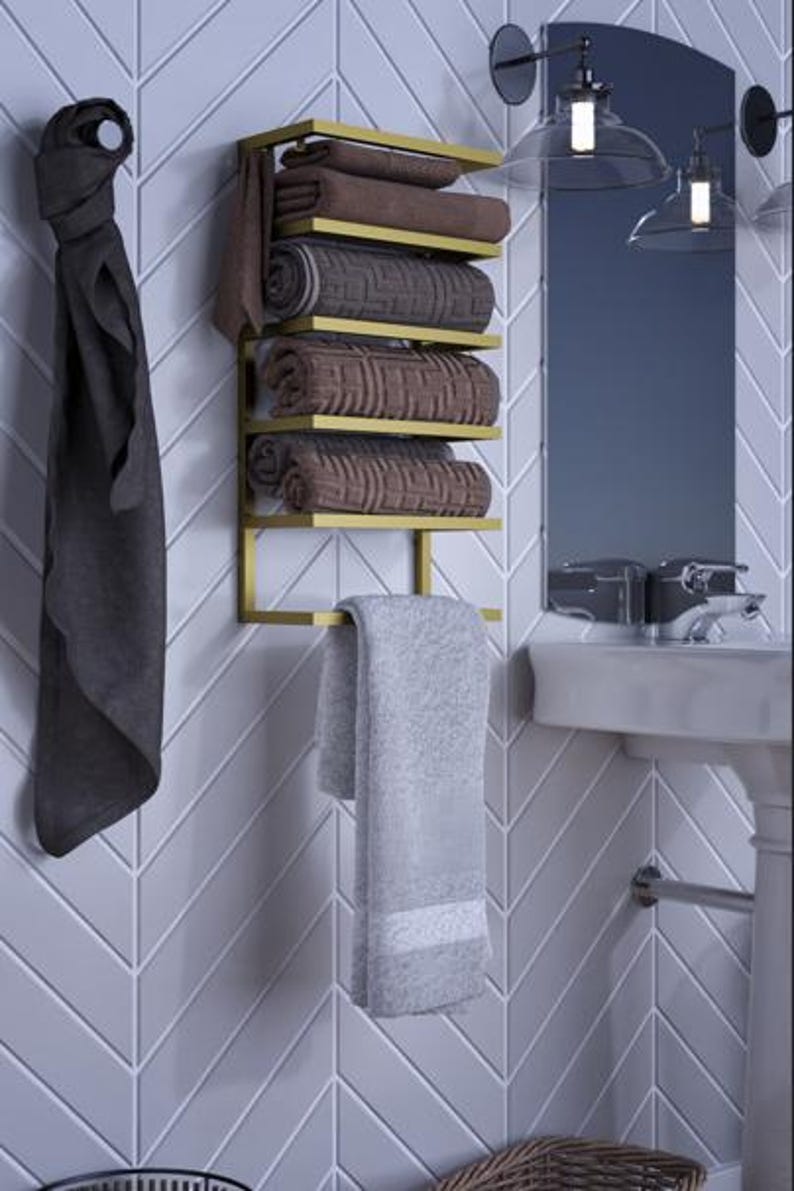 May include: A gold-colored metal towel rack with five shelves, holding rolled brown towels. A gray towel hangs from the bottom. A dark gray towel hangs on the left. The rack is mounted on a white, chevron-patterned wall.