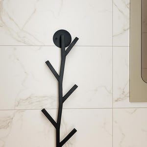 May include: Black metal wall-mounted coat rack in a tree branch design. The rack has a circular mounting plate and several angled branches for hanging items. It is mounted on a white tiled wall.