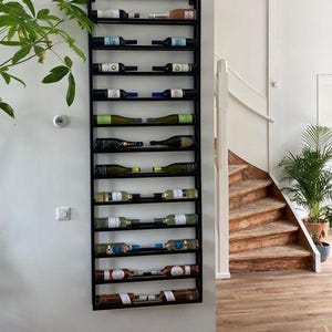 May include: A black metal wine rack mounted on a white wall, holding various wine bottles. The rack has multiple horizontal shelves, displaying bottles of different colors and labels. A staircase and potted plants are visible in the background.