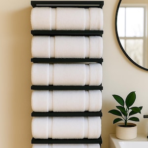 May include: A black, wall-mounted towel rack holding six rolled white towels. The rack is made of black metal and is mounted on a light beige wall. A round black mirror and a small potted plant are also visible.