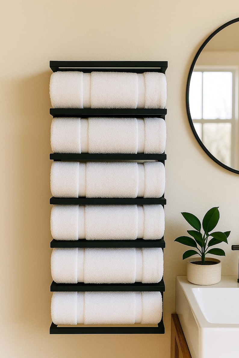 May include: A black, wall-mounted towel rack holding six rolled white towels. The rack is made of black metal and is mounted on a light beige wall. A round black mirror and a small potted plant are also visible.