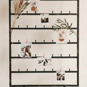 May include: Black metal photo display with horizontal bars and small pegs for hanging photos. Several photos are clipped to the bars with small wooden clothespins. The display is decorated with sprigs of greenery and fairy lights.