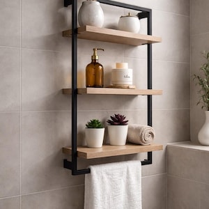 May include: A wall-mounted bathroom shelf unit with three wooden shelves and a black metal frame. The shelves hold decorative items like a vase with dried plants, a candle, and small potted succulents. A white towel hangs from the bottom shelf.
