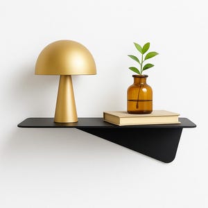 Puede incluir: Estantería de pared negra moderna con una lámpara dorada en forma de hongo, un pequeño jarrón de cristal ámbar con hojas verdes y un libro cerrado. La estantería tiene un diseño minimalista y está montada en una pared blanca.
