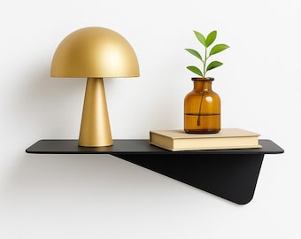 May include: A modern black wall shelf with a gold mushroom-shaped lamp, a small amber glass vase with green leaves, and a closed book. The shelf has a minimalist design and is mounted on a white wall.