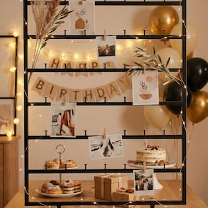Könnte beinhalten: Ein schwarzer Metall-Fotoständer mit einem "Happy Birthday"-Banner, Fotos und dekorativen Elementen. Der Ständer hält Fotos, die mit Holzklammern befestigt sind, mit Kuchen, Geschenken und Luftballons. Lichterketten sorgen für einen warmen Schein.