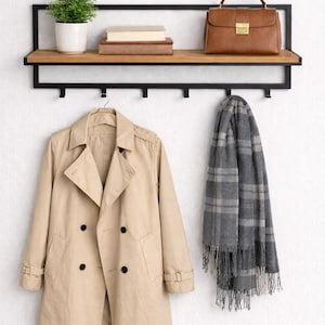 Könnte beinhalten: Wandregal mit schwarzem Metallrahmen und Holzregal. Ein beigefarbener Trenchcoat hängt darunter, mit einem grau karierten Schal und einer braunen Lederhandtasche auf dem Regal. Eine kleine Topfpflanze und Bücher befinden sich ebenfalls auf dem Regal.