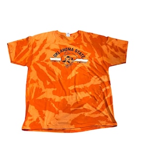 Peut inclure: T-shirt orange tie-dye avec un logo noir et blanc Oklahoma State représentant un mascotte de cowboy. Le logo indique "Oklahoma State" avec "Stillwater, OK" et "#GoPokes" en texte plus petit.