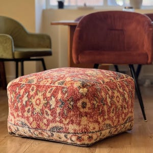 May include: A square, upholstered ottoman with a detailed, vintage-style pattern in red, beige, and black. The ottoman is placed on a hardwood floor, with a blurred background featuring chairs and a window.
