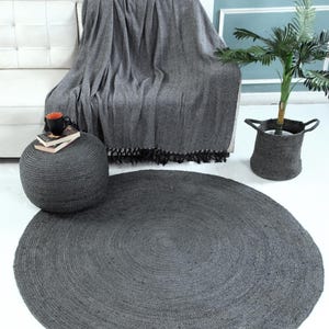 May include: A round, dark gray jute rug with a woven texture. The rug is placed on a white floor in a living room setting.