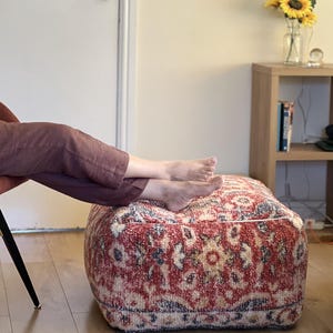 Może przedstawiać: Kwadratowa pufa we wzory z czerwono-kremowym kwiatowym wzorem. Pufa to niskie, amortyzowane siedzisko, idealne do salonu lub sypialni. Projekt obejmuje odcienie czerwieni, kremu i niebieskiego. Pufa ma około 46 cm kwadratowych.