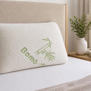 以下が含まれることがあります： 緑色の竹の葉のデザインと「Bamboo」の文字がプリントされた、長方形のオフホワイトの竹枕。枕は白いベッドの上にあります。