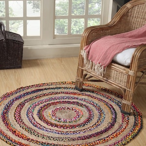 May include: A round braided jute rug with concentric circles of natural, beige, and colorful threads. The rug is on a wooden floor, near a wicker chair with a pink blanket and a dark brown basket.