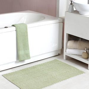 May include: A light green rectangular bath mat with a white chevron pattern on a white floor. A matching green towel hangs on the edge of a white bathtub. The bathroom also features a white sink and shelving.
