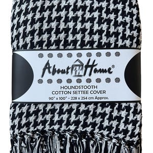 Op de afbeelding: Een zwart-witte bankhoes van katoen met een pied-de-poule patroon en franjes. Een papieren band om het midden bevat de tekst About Home, HOUNDSTOOTH COTTON SETTEE COVER en de afmetingen 228 cm bij 254 cm.