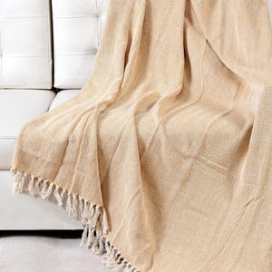 May include: A soft, beige throw blanket with a subtle herringbone pattern and fringe trim. The blanket is draped over a white couch.