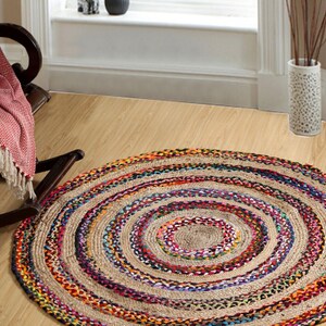 May include: A round jute rug with concentric circles of natural beige and colorful braided accents. The rug features a central beige area surrounded by rings of vibrant colors like purple, blue, and orange. It is placed on a light wood floor.