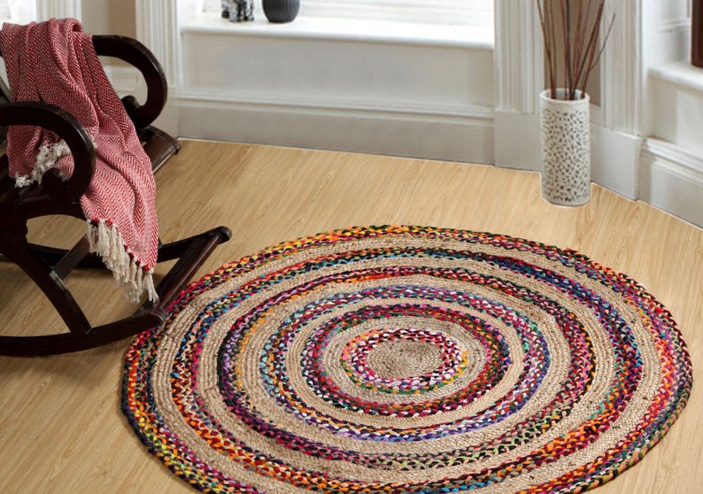 May include: A round jute rug with concentric circles of natural beige and colorful braided accents. The rug features a central beige area surrounded by rings of vibrant colors like purple, blue, and orange. It is placed on a light wood floor.