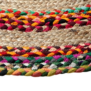 May include: Close-up of a round jute rug with colorful braided fabric accents. The rug features a natural beige woven base with bands of red, orange, yellow, green, and purple fabric. The braided sections add texture and visual interest to the floor covering.