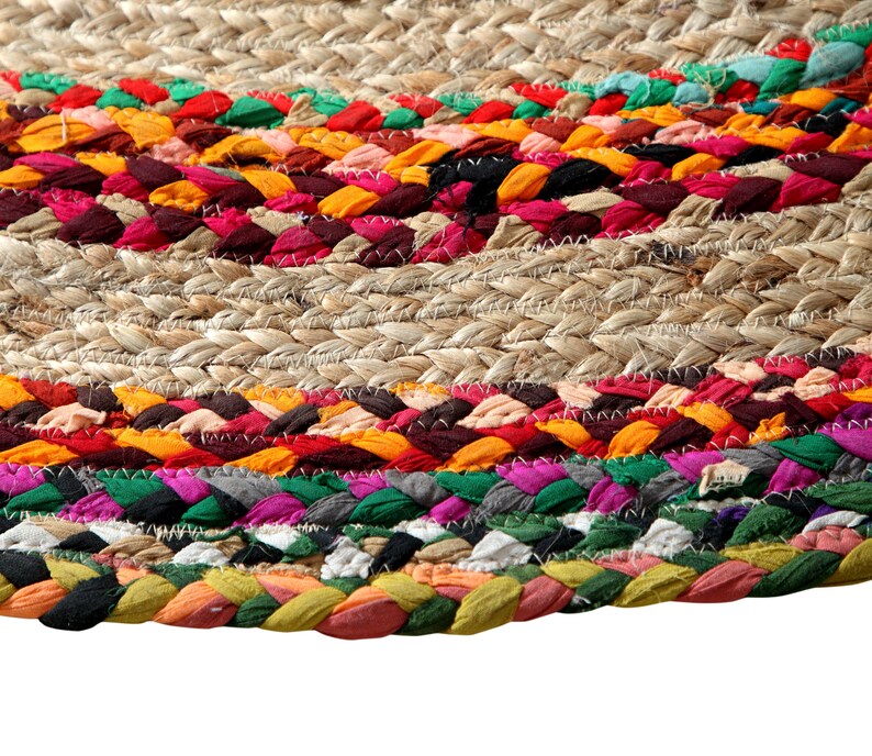 May include: Close-up of a round jute rug with colorful braided fabric accents. The rug features a natural beige woven base with bands of red, orange, yellow, green, and purple fabric. The braided sections add texture and visual interest to the floor covering.