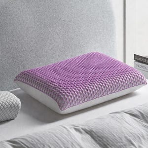 May include: A lavender pillow with a honeycomb-patterned top and white sides rests on a bed. A smaller, gray pillow with a diamond pattern is also visible. The scene includes a gray headboard and a stack of books.