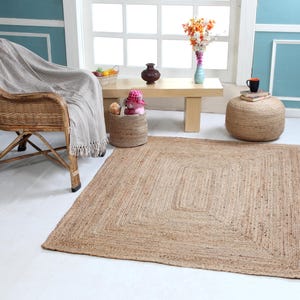 Op de afbeelding: Een natuurlijk jute vloerkleed met een geweven ontwerp in een lichtbruine kleur. Het vloerkleed is geplaatst in een woonkamer met een rieten stoel, een houten tafel en een geweven opbergmand.