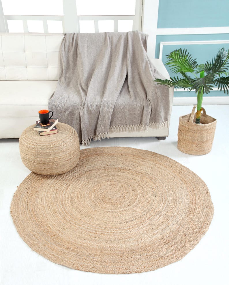 May include: A round, natural jute rug with a woven texture. The rug is light brown and has a circular shape. It is placed on a white floor in a living room setting.