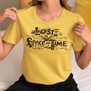 Könnte beinhalten: Senfgelbes T-Shirt mit dem schwarzen Schriftzug "Lost in Space-Time". Das Design beinhaltet eine Sanduhr, mathematische Symbole und die Gleichung E=mc². Das T-Shirt ist aus weichem Material.