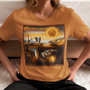 May include: Mustard-colored t-shirt featuring a surrealist painting with the word "Mercury" at the top. The artwork depicts melting clocks, a bright sun, and a landscape. The shirt is short-sleeved and has a crew neck.