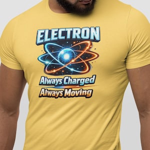 Könnte beinhalten: Senfgelbes T-Shirt mit einer Atomgrafik und dem Wort "ELECTRON". Unter der Atomgrafik stehen die Worte "Always Charged Always Moving" in Blau und Orange. Ein Design zum Thema Wissenschaft.