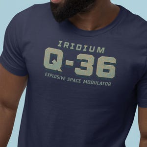 May include: Navy blue t-shirt with the text "IRIDIUM Q-36 EXPLOSIVE SPACE MODULATOR" in a distressed, vintage style. The text is in a light teal color, and the shirt has a crew neck.