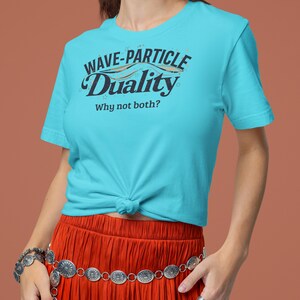 Könnte beinhalten: Ein türkisfarbenes T-Shirt mit dem Text "WAVE-PARTICLE DUALITY" und "Why not both?" in Schwarz und Weiß. Das T-Shirt ist an der Taille geknotet. Die Person trägt einen roten Rock und Silberschmuck.