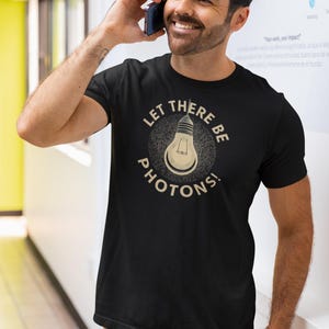 Pode incluir: Camiseta preta com o texto "LET THERE BE PHOTONS!" e um gráfico de lâmpada. A camiseta é usada por uma pessoa. A pessoa está sorrindo e segurando um telefone na orelha. A pessoa está usando calças marrons.
