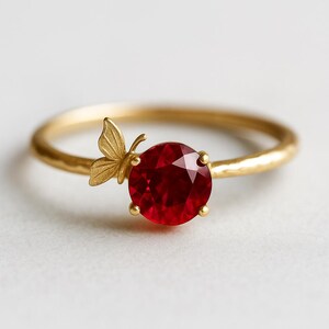 Op de afbeelding: Een gouden ring met een grote, ronde, dieprode edelsteen. Aan de zijkant van de edelsteen zit een klein vlinderdetail. De ring heeft een getextureerde band. De achtergrond is wit.