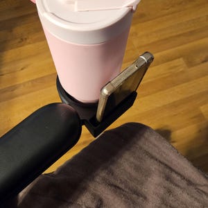 May include: A pink insulated tumbler with a straw and a black phone holder attached. The tumbler has a white lid with a pink flip-top closure. A smartphone is in the phone holder. The tumbler is on a wooden surface.