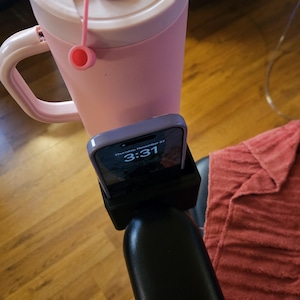 May include: A pink insulated tumbler with a handle and a straw, next to a phone in a black holder. The phone displays the time as 3:31. The tumbler has a lid with a small tab.