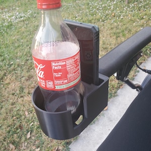 May include: A black plastic cup holder and phone mount attachment secured to a black armrest. The cup holder contains a clear plastic bottle with a red label and white text, while the adjacent rectangular slot holds a black smartphone upright.