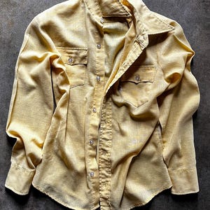 May include: A light yellow button-down shirt with two chest pockets and long sleeves. The shirt has a classic collar and is made of a textured fabric. The shirt is unbuttoned, and the fabric has a slightly wrinkled appearance.