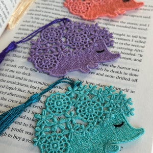May include: Three handmade fabric bookmarks shaped like hedgehogs. The bookmarks are made of delicate lace and feature a variety of colors, including purple, pink, and teal.