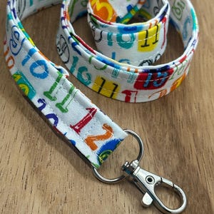 May include: A white lanyard with a colorful number pattern. The lanyard has a silver-colored clasp and a metal ring. The numbers are in various colors, including blue, green, red, and yellow.