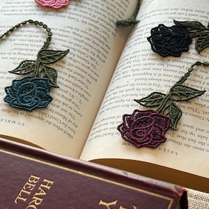 May include: A book with a bookmark made of delicate lace flowers in shades of pink, teal, black, and burgundy. The bookmark is made of several flowers connected by a chain.