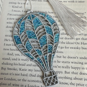 May include: A silver and blue hot air balloon bookmark with a white tassel. The bookmark is made of fabric and has a delicate, intricate design.