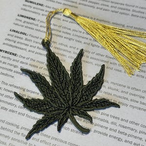 May include: A green embroidered marijuana leaf bookmark with gold tassels. The bookmark is laying on a page of a book with text about terpenes and their properties.