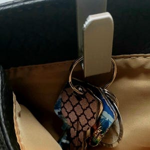 Pode incluir: Um gancho de bolsa prateado está preso à borda de uma bolsa preta. Um porta-chaves com várias chaves está pendurado no gancho. As chaves têm desenhos decorativos.