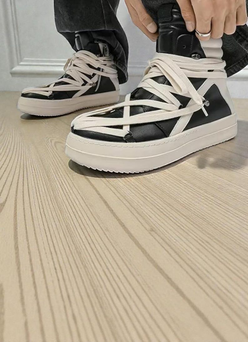 Pode incluir: T&ecirc;nis de cano alto preto e branco com cadar&ccedil;os creme. Os t&ecirc;nis t&ecirc;m uma sola grossa, branca suja e um design de cano alto. Os cadar&ccedil;os envolvem o tornozelo e a parte superior do sapato.