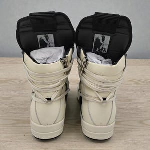 Pode incluir: Par de t&ecirc;nis de cano alto creme e preto. Os t&ecirc;nis apresentam fecho de z&iacute;per, cadar&ccedil;os enrolados e uma l&iacute;ngua preta acolchoada. O design inclui um logotipo na l&iacute;ngua e uma sola grossa.