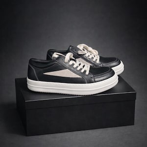 May include: Black leather sneakers with white soles and laces. The shoes feature a light-colored triangle design on the sides. The sneakers are displayed on a black box against a dark background. A stylish and modern footwear option.