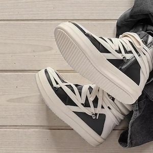 Pode incluir: T&eacute;nis de cano alto em preto e branco. Os sapatos apresentam uma parte superior em couro preto, atacadores brancos e uma sola grossa branca. Um detalhe de z&iacute;per &eacute; vis&iacute;vel na lateral. Colocados sobre uma superf&iacute;cie de madeira clara.